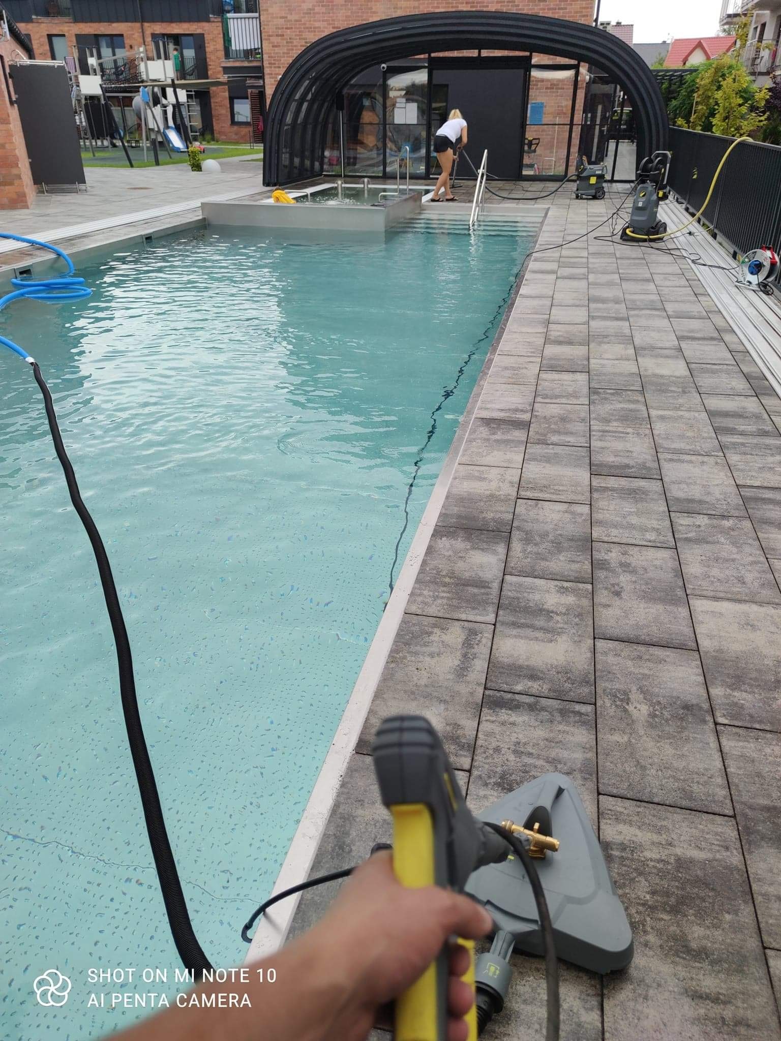 Czyszczenie kostki brukowej wokół basenu zewnętrznego przy użyciu myjki ciśnieniowej z żółtymi akcentami, widok z perspektywy osoby trzymającej dyszę, w tle osoba czyszcząca basen.