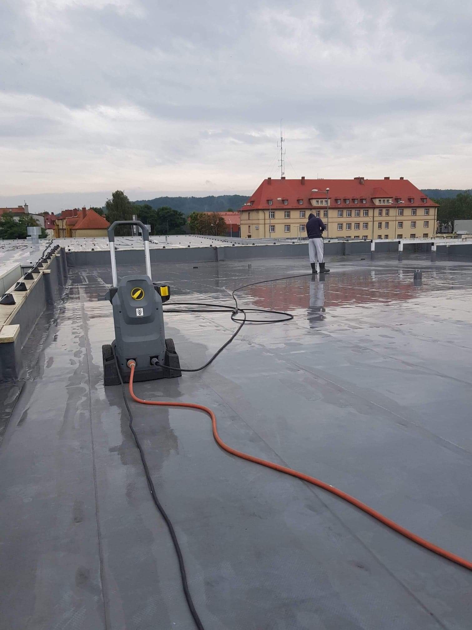 Czyszczenie dachu płaskiego za pomocą myjki ciśnieniowej; widoczny pracownik w kombinezonie, duży budynek w tle.