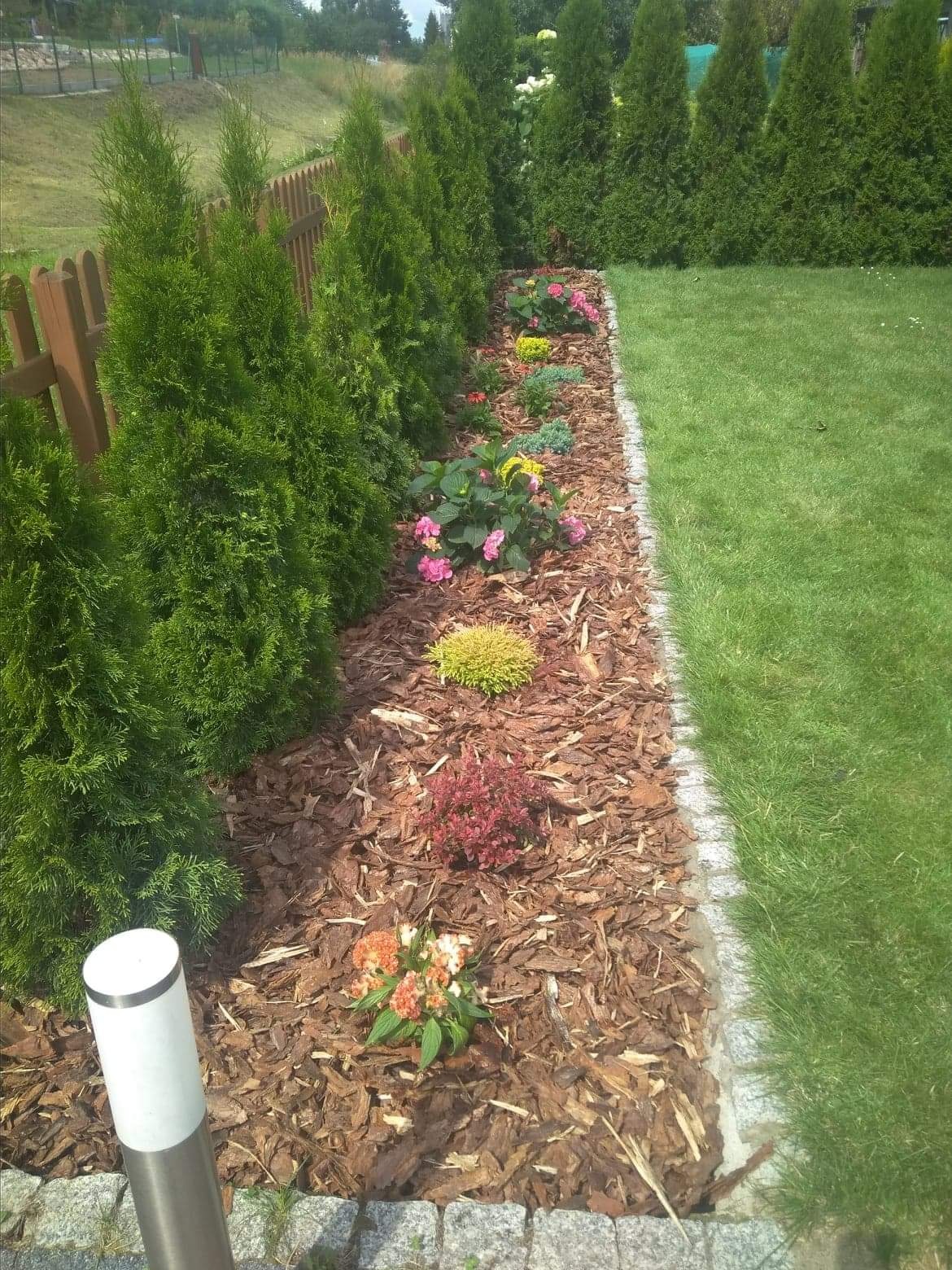 Rabata z korą, kwiatami i iglakami wzdłuż ogrodzenia, z trawnikiem po prawej stronie, oświetlona lampą ogrodową