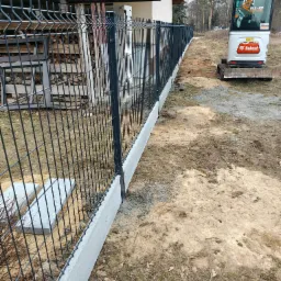 Nowoczesne ogrodzenie panelowe z podmurówką betonową, widoczne z perspektywy ukośnej. W tle mini koparka Bobcat i stos drewna. Elementy metalowe w kolorze grafitowym.