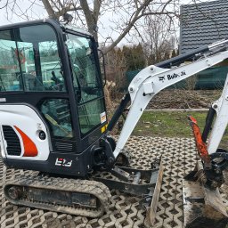 WILK - KOP ROBOTY ZIEMNE USŁUGI MINIKOPARKĄ PAWEŁ WILK - Mini koparka Bobcat na kostce brukowej. Widoczny łyżka koparki, gąsienice i kabina. W tle dom i drzewa. Sprzęt budowlany gotowy do pracy.