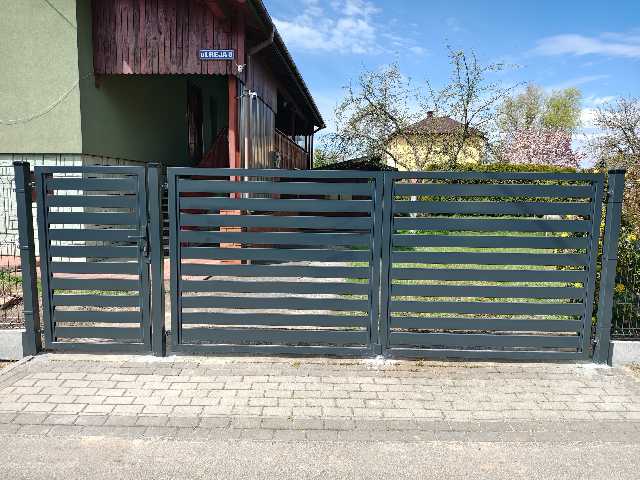 Nowoczesne, poziome ogrodzenie panelowe w kolorze grafitowym z furtką i bramą wjazdową na posesji z kostką brukową, widok z zewnątrz.