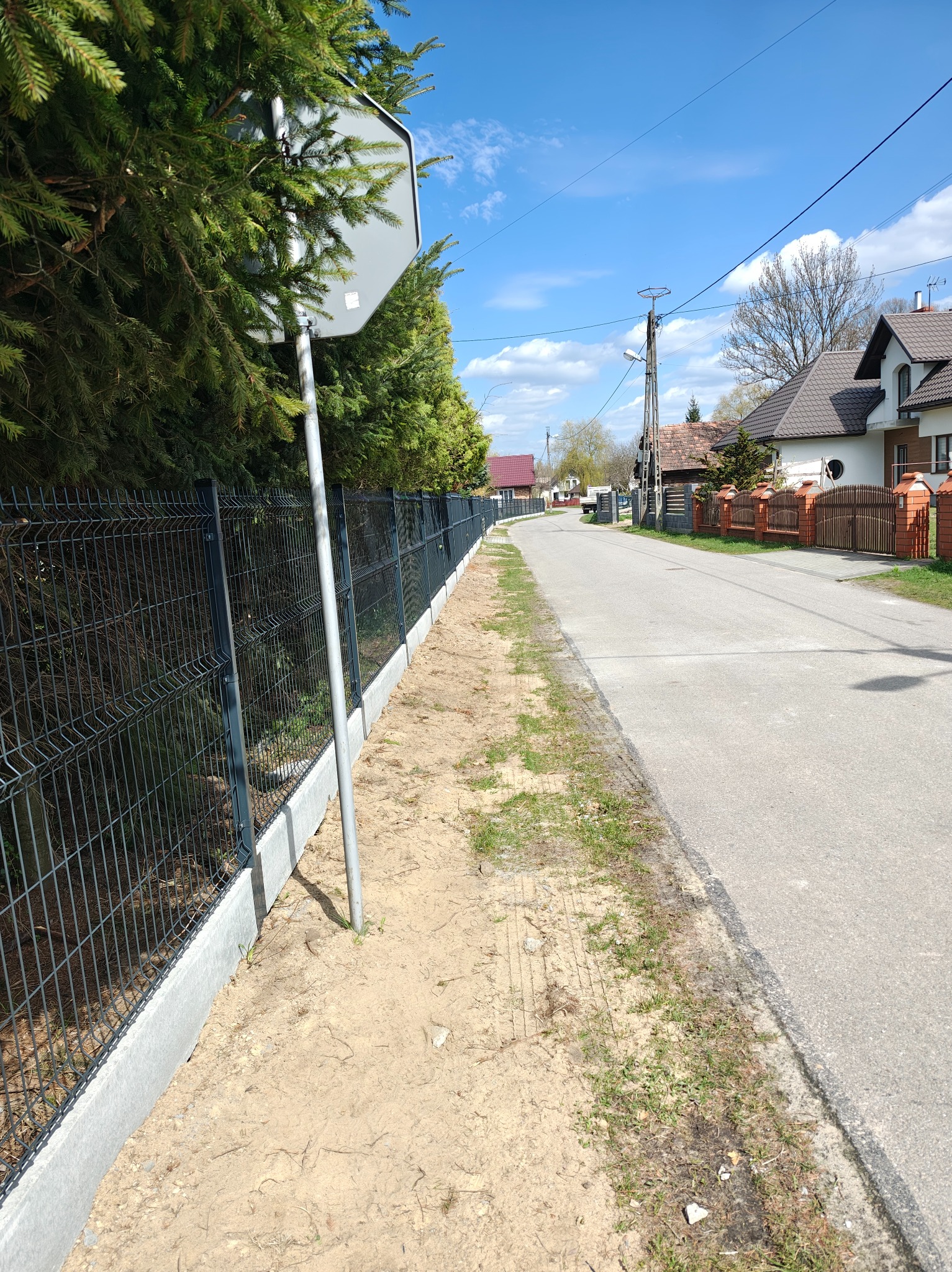 Nowoczesne, metalowe ogrodzenie panelowe z podmurówką betonową, ciągnące się wzdłuż drogi, z zielonymi tujami w tle i błękitnym niebem.