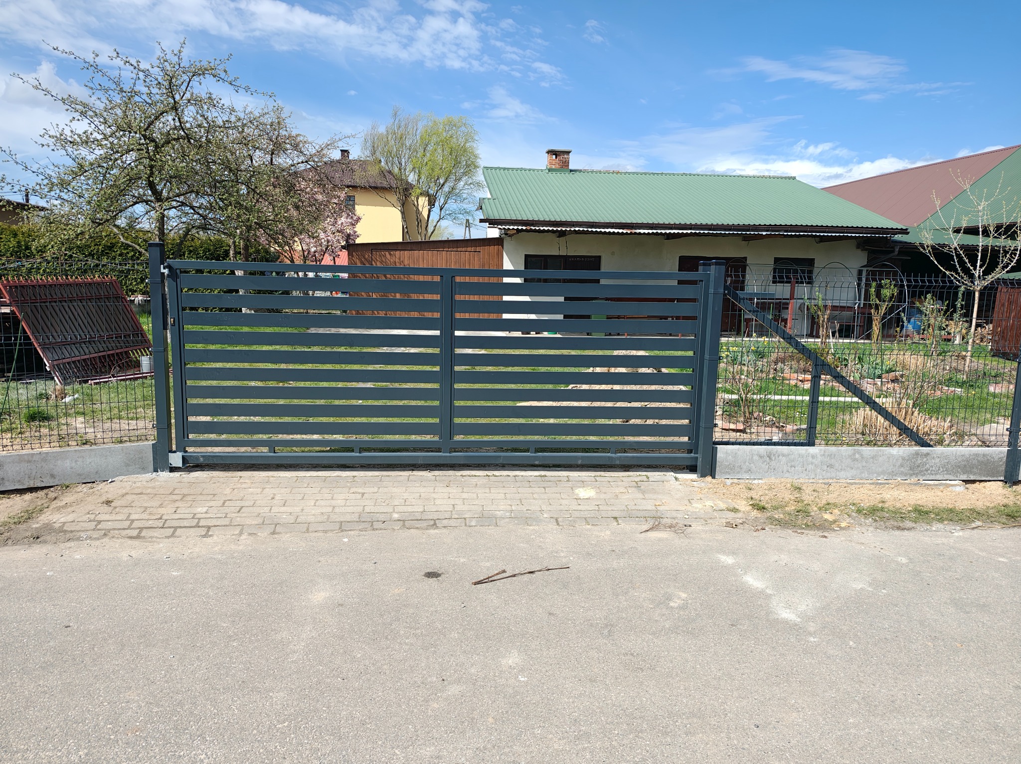 Nowoczesna, przesuwna brama w kolorze grafitowym z poziomymi panelami, zamontowana na posesji z kostką brukową i widocznym domem w tle.