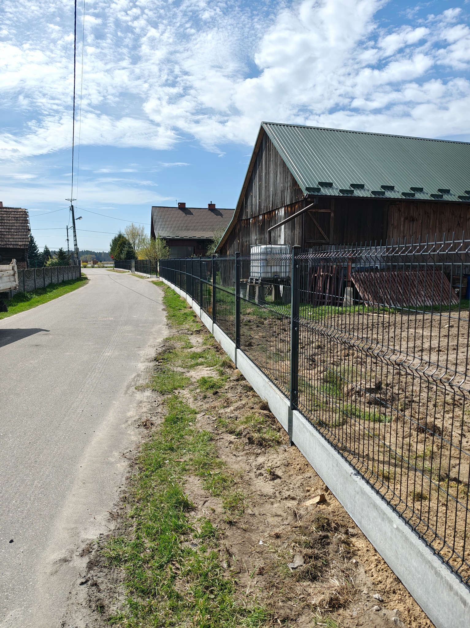 Nowoczesne ogrodzenie panelowe z podmurówką betonową, oddzielające posesję od drogi wiejskiej. W tle zabudowania gospodarcze i błękitne niebo z chmurami.