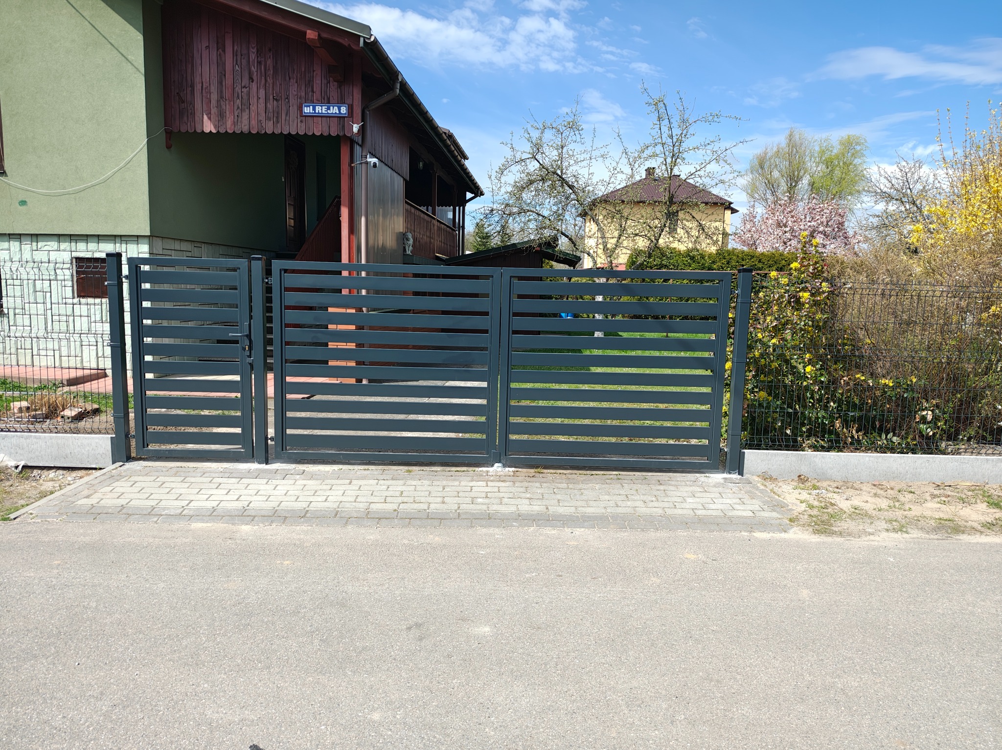 Nowoczesna, pozioma brama wjazdowa w kolorze grafitowym, z widoczną kostką brukową u podstawy i domem w tle, ul. Reja 8.