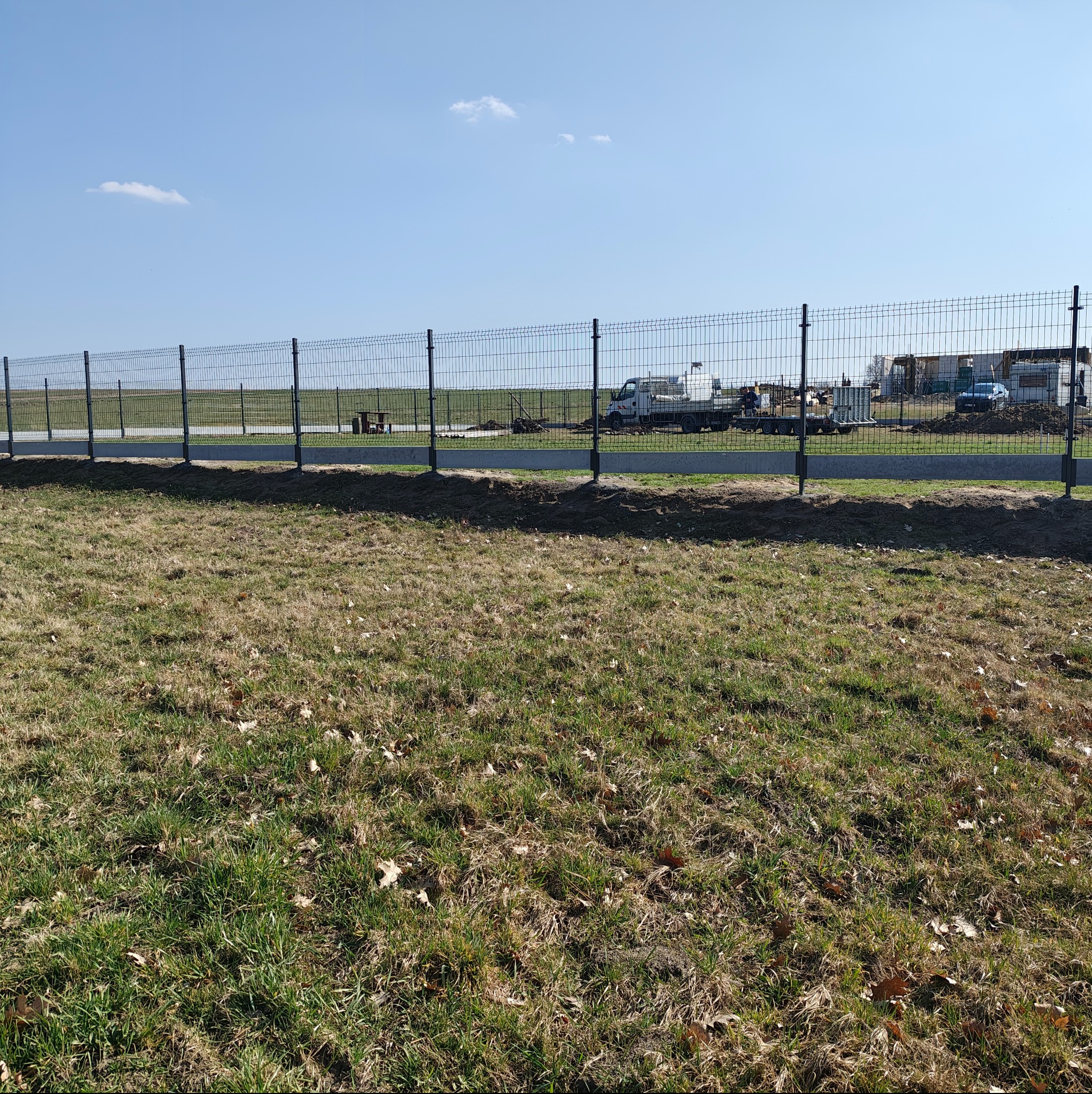 Długi, panelowy, metalowy płot z podmurówką, oddzielający teren zielony od placu budowy z zaparkowanymi pojazdami w tle, pod błękitnym niebem.