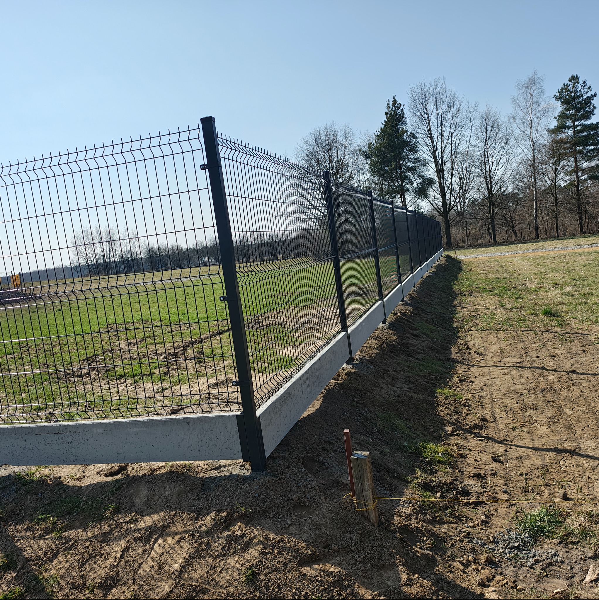 Nowoczesne, panelowe ogrodzenie z podmurówką betonową, widok z boku na tle zieleni i drzew. Słupki w kolorze grafitowym. Widoczne ślady prac ziemnych.
