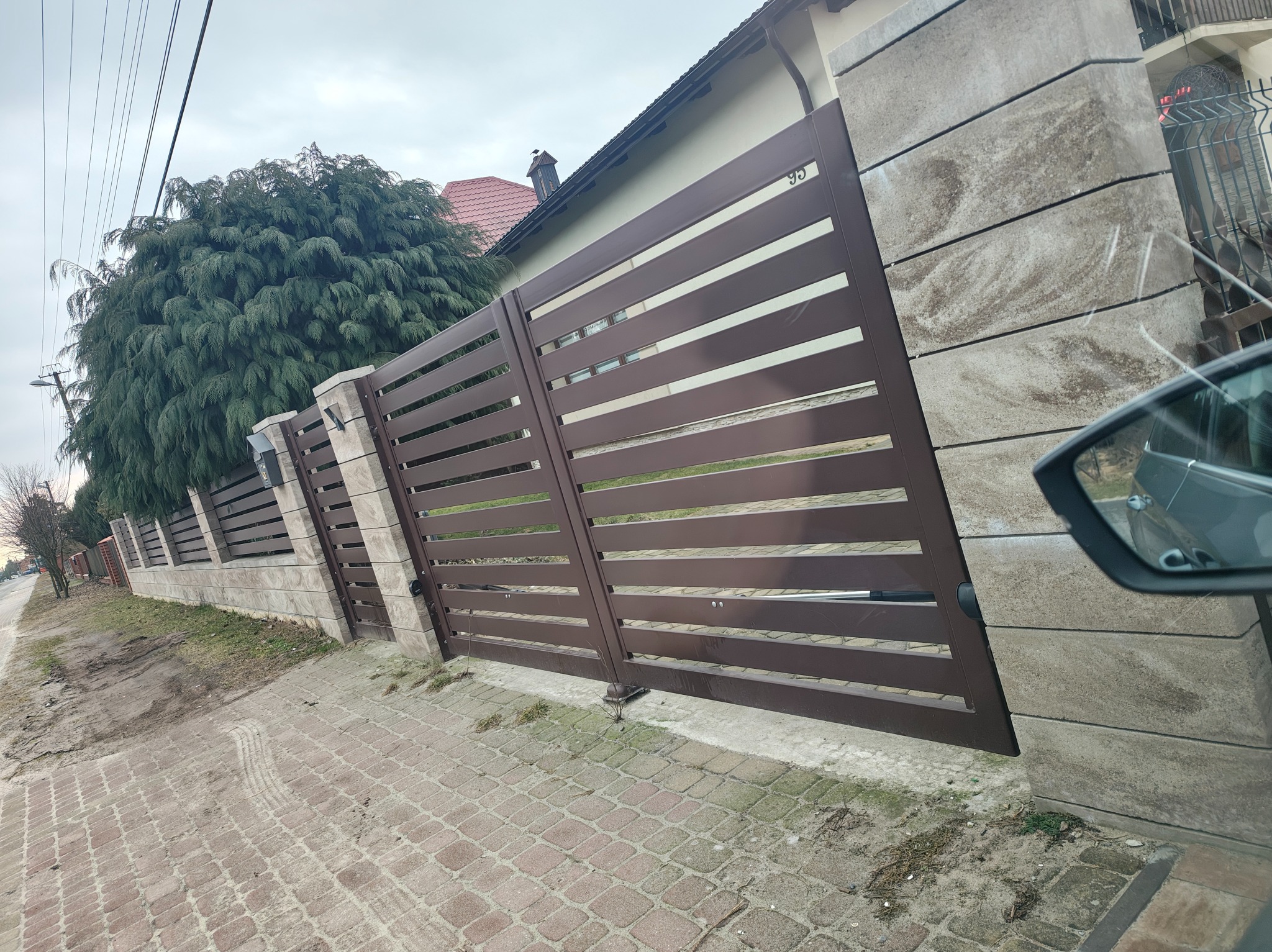 Nowoczesne ogrodzenie panelowe w kolorze brązowym z betonowymi słupkami, widoczne z perspektywy chodnika. Widoczny fragment samochodu i zieleń w tle.