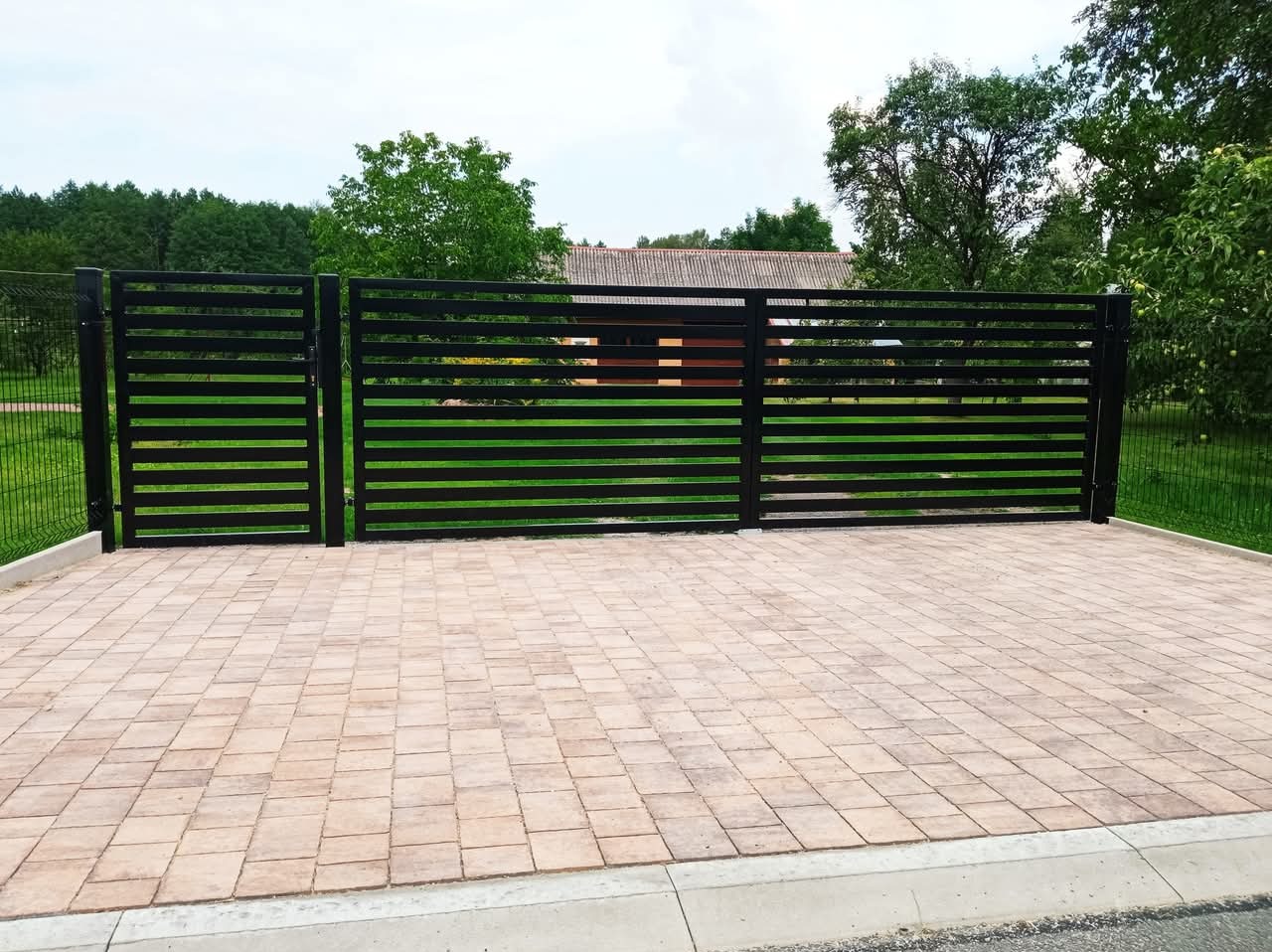 Nowoczesne, czarne ogrodzenie panelowe z poziomymi szczeblami, widok z zewnątrz na posesję wyłożoną kostką brukową, w tle zieleń drzew i budynek.