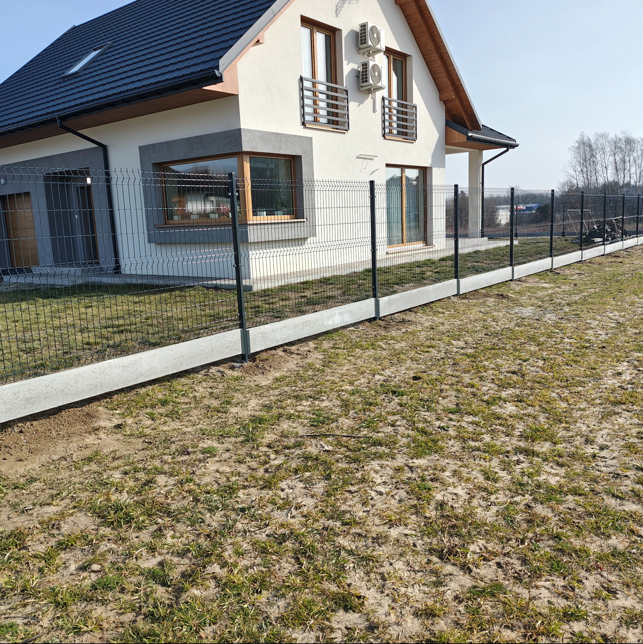 Nowoczesne ogrodzenie panelowe, czarne, z podmurówką betonową, na tle jasnego domu z dwoma oknami i klimatyzacją na elewacji. Widok z perspektywy trawnika.