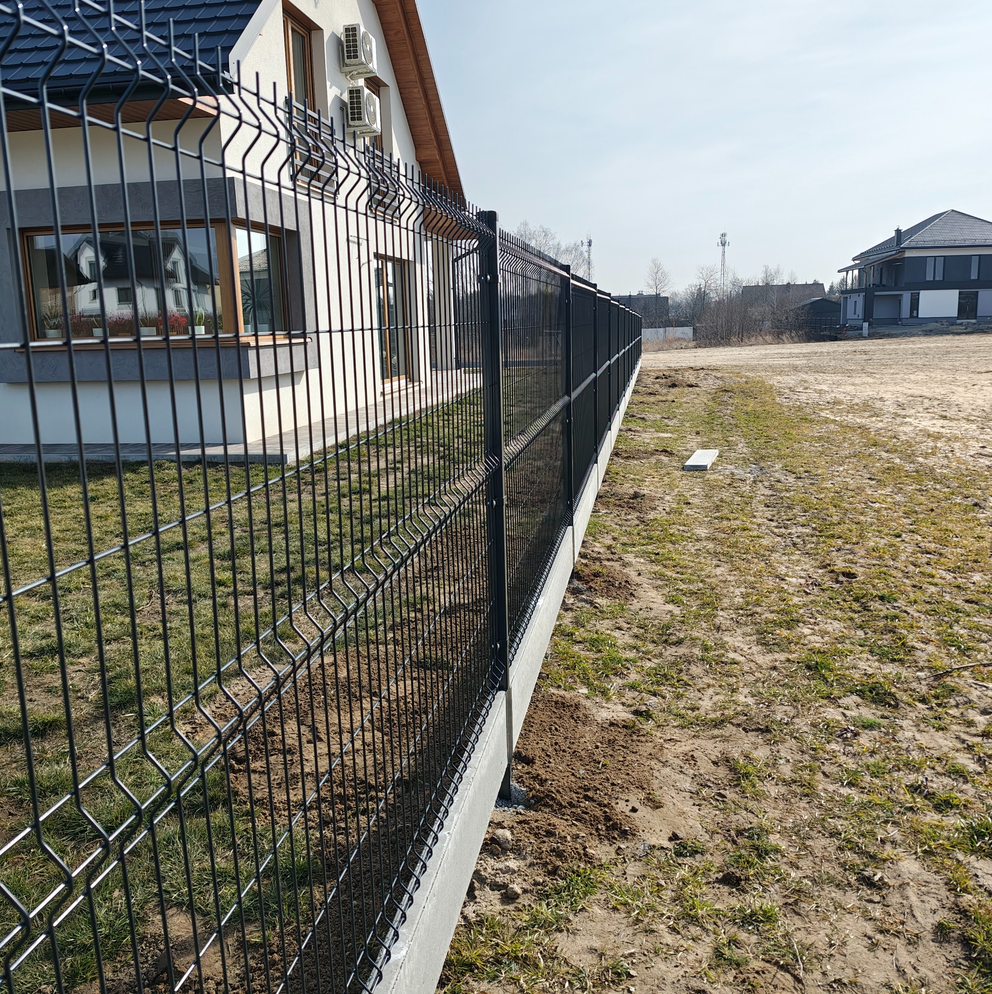 Czarny, panelowy płot na betonowej podmurówce, oddzielający posesję z domem o białej elewacji i szarym dachem, widok z perspektywy ziemi, słoneczny dzień.