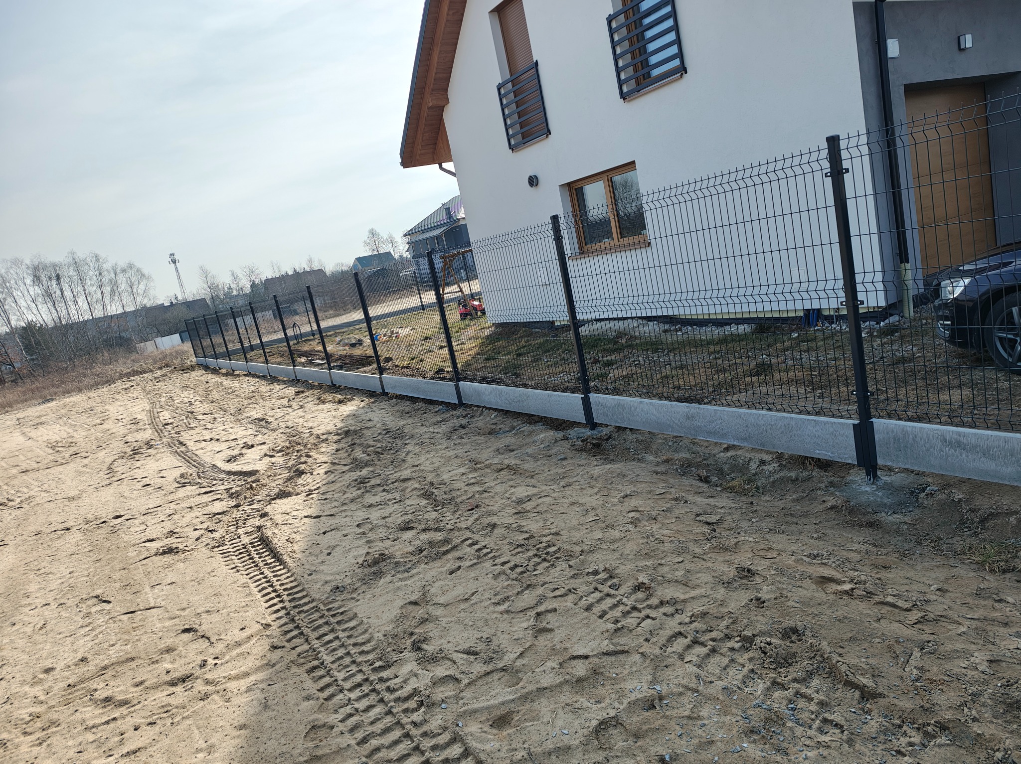 Nowoczesne ogrodzenie panelowe z podmurówką betonową, otaczające posesję z białym domem. Ślady opon na piaszczystym terenie. Słoneczny dzień.