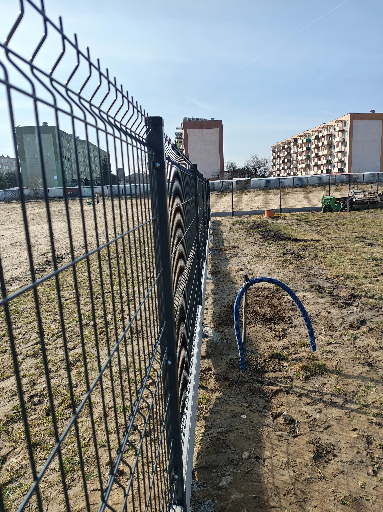 Nowoczesne, grafitowe ogrodzenie panelowe na nierównym terenie. W tle bloki mieszkalne i błękitne niebo. Widoczne przygotowania do dalszych prac ziemnych.