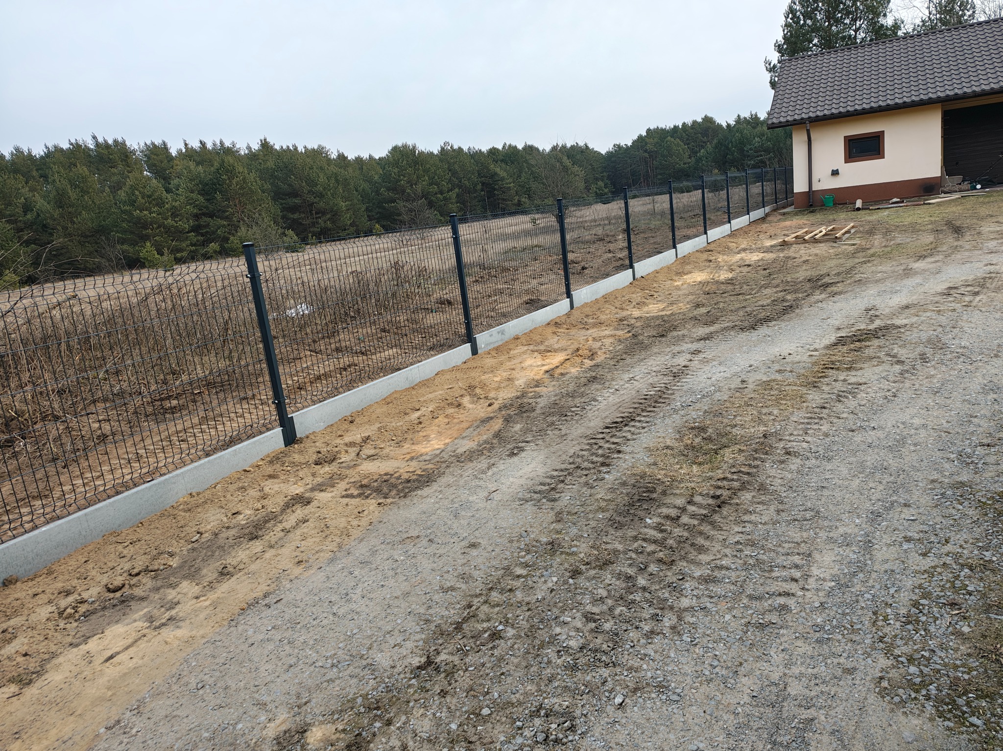 Nowoczesne ogrodzenie panelowe z podmurówką betonową, widok z boku na tle lasu i budynku gospodarczego, szara kolorystyka. Świeżo wyrównany teren.