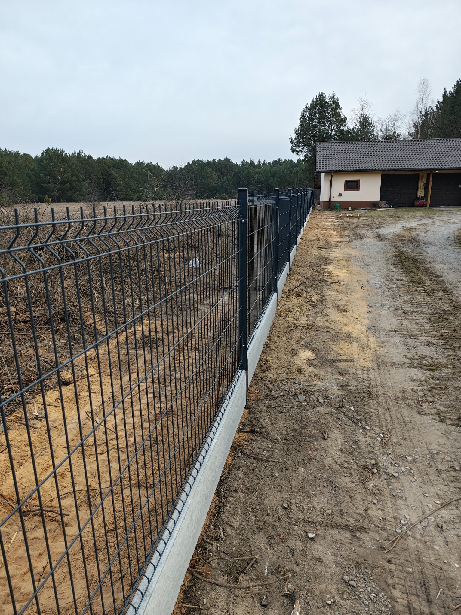 Nowoczesne, panelowe ogrodzenie z podmurówką betonową, w tle dom jednorodzinny i las. Widok na świeżo zamontowane ogrodzenie w surowym otoczeniu.