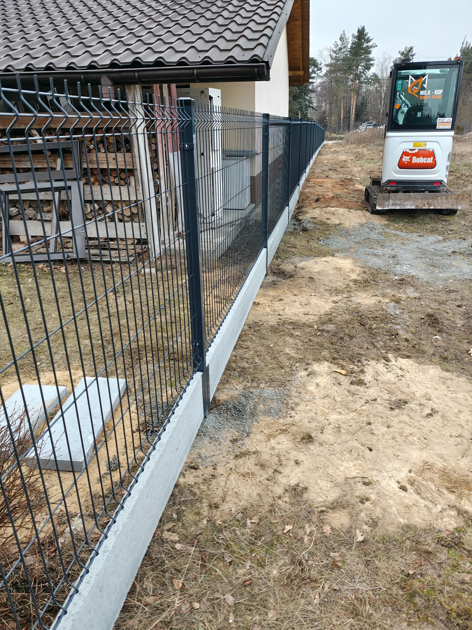 Nowoczesne ogrodzenie panelowe z podmurówką betonową, widoczne z perspektywy ukośnej. W tle mini koparka Bobcat i stos drewna. Elementy metalowe w kolorze grafitowym.