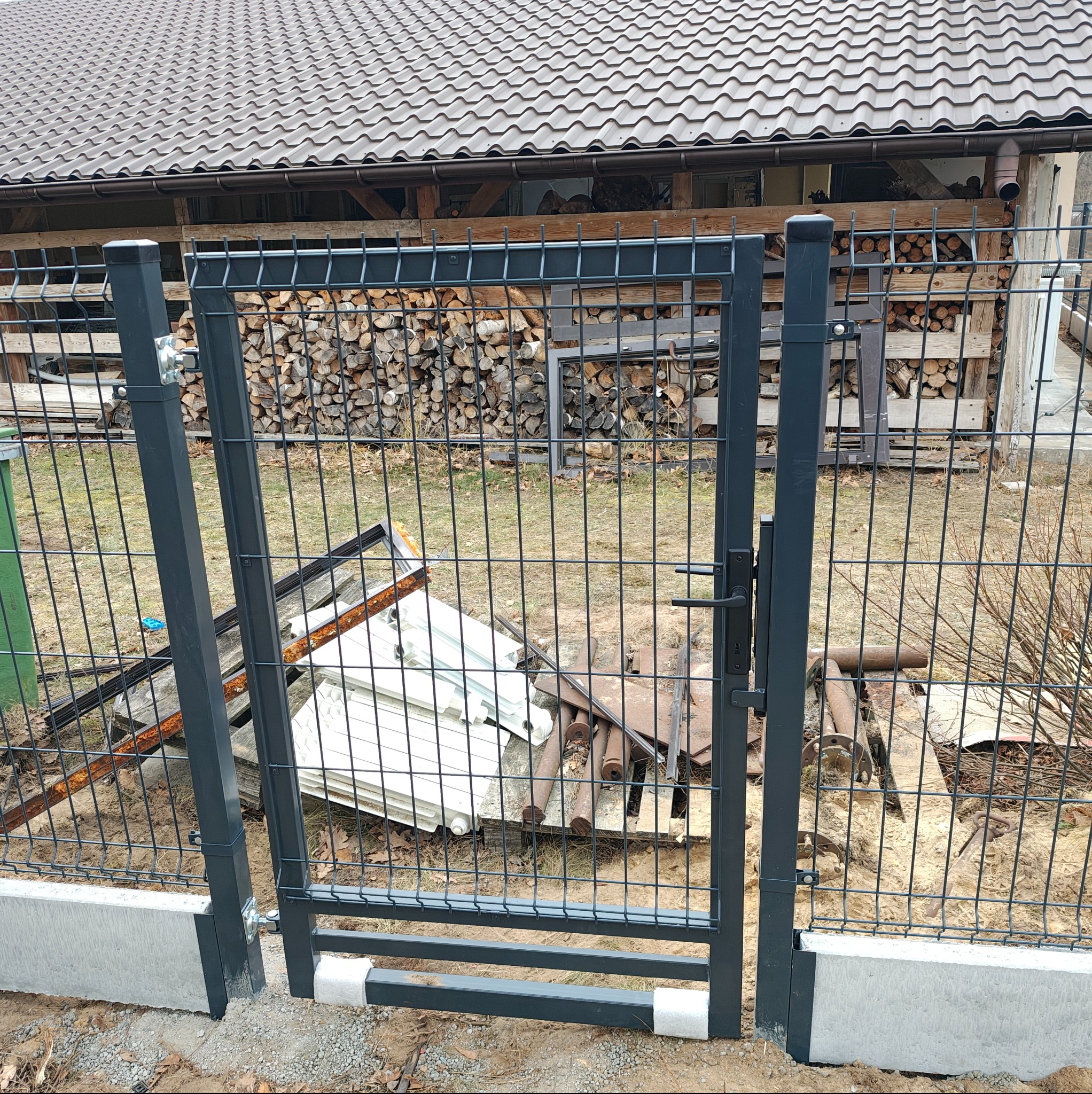 Nowa, grafitowa furtka z siatki panelowej, zamontowana w Rudniku nad Sanem. W tle sterta drewna i elementy starego ogrodzenia.