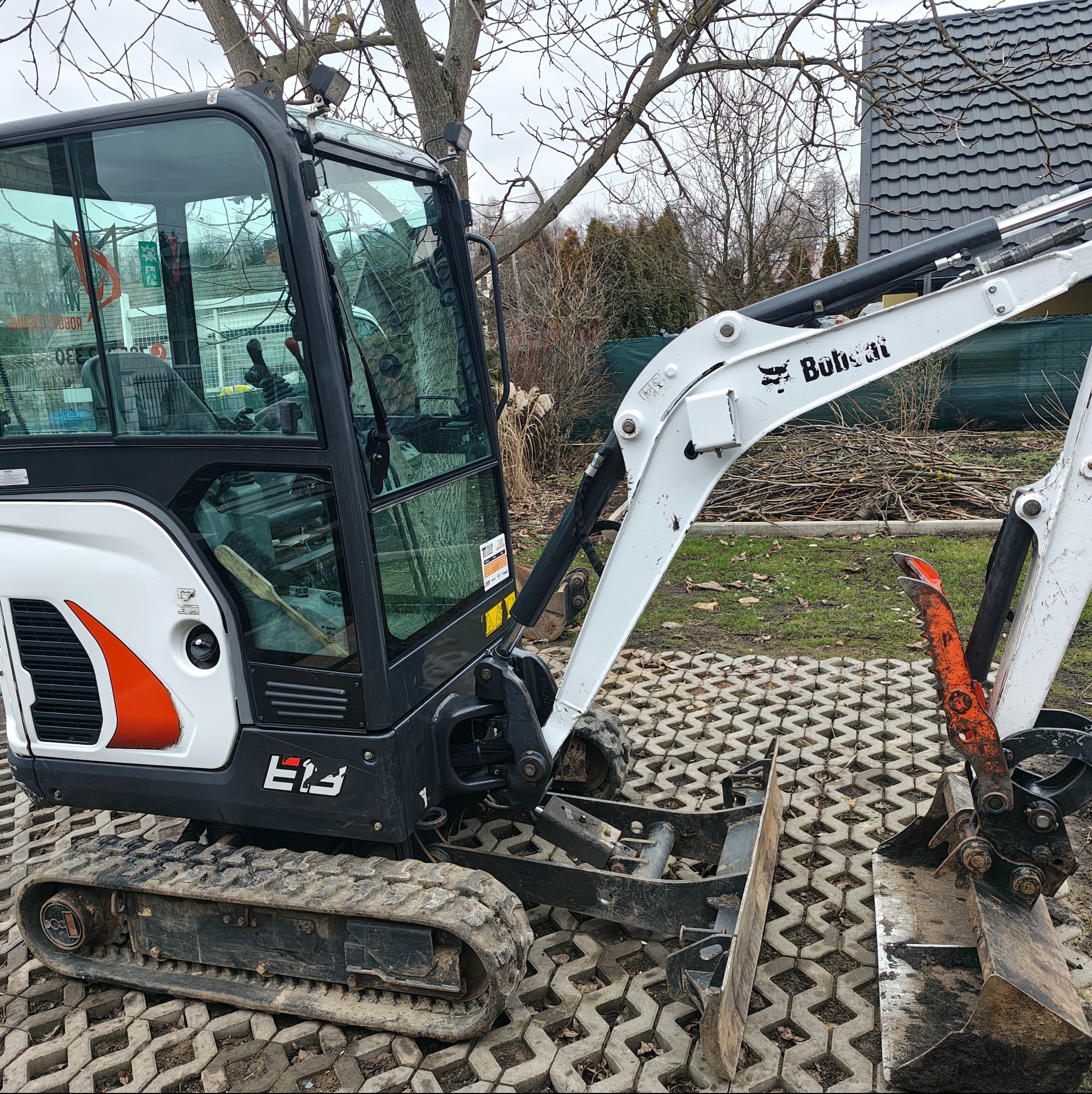 Mini koparka Bobcat na kostce brukowej. Widoczny łyżka koparki, gąsienice i kabina. W tle dom i drzewa. Sprzęt budowlany gotowy do pracy.