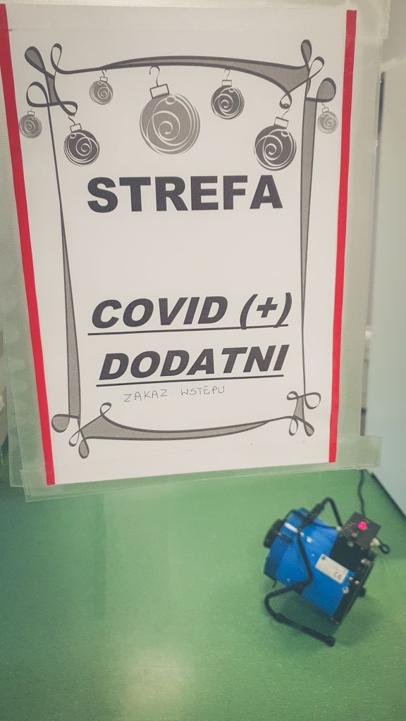 Niebieski osuszacz powietrza ustawiony na zielonej podłodze obok kartki z napisem 'Strefa Covid (+) Dodatni, Zakaz Wstępu' ozdobionej motywem świątecznym.