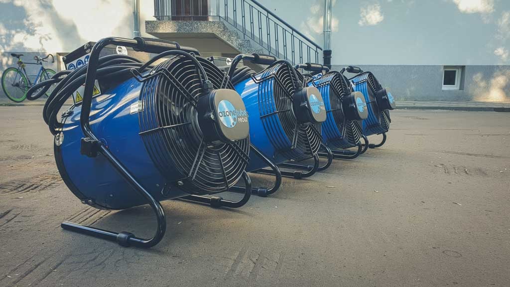 Szereg niebieskich wentylatorów przemysłowych z czarnymi obudowami i napisem 'Ozonetubes PRO42' ustawionych na zewnątrz, z rowerem opartym o ścianę w tle.