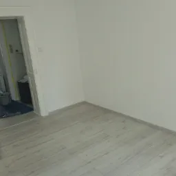 Jasne panele podłogowe imitujące drewno w nowo wyremontowanym pokoju w Pasymiu, widoczne przygotowania do malowania w sąsiednim pomieszczeniu.