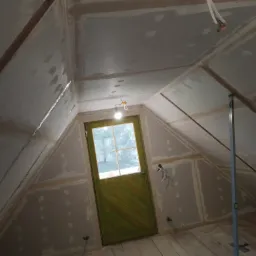 Poddasze w trakcie wykańczania: płyty karton-gips na skosach i suficie, szpachlowane łączenia, drewniana podłoga, zielone drzwi ze szklanym oknem, widoczne przewody elektryczne i poziomica.
