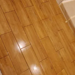 Ułożone, błyszczące, podłużne płytki podłogowe imitujące drewno w odcieniach brązu, widoczne krzyżyki dystansowe, fragment ściany z taśmą malarską.