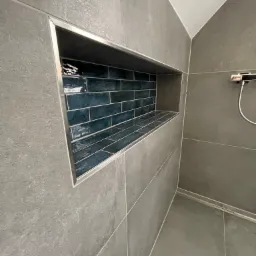 Wnęka w ścianie kabiny prysznicowej wyłożona ciemnoturkusową, błyszczącą mozaiką cegiełkową z jasną fugą, obramowana metalową listwą, na tle szarych, matowych płytek wielkoformatowych.