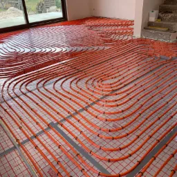 Pomarańczowe rury ogrzewania podłogowego ułożone na srebrnej folii refleksyjnej z czarnymi mocowaniami, widoczne fragmenty ścian i schodów w surowym stanie.