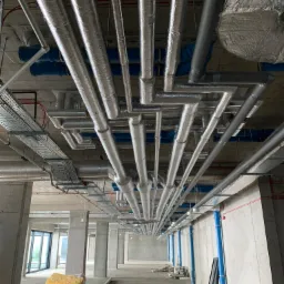 Widok z dołu na system rur wentylacyjnych i instalacyjnych pod sufitem w surowym stanie, z widoczną izolacją i elementami mocującymi w nowo budowanym obiekcie.