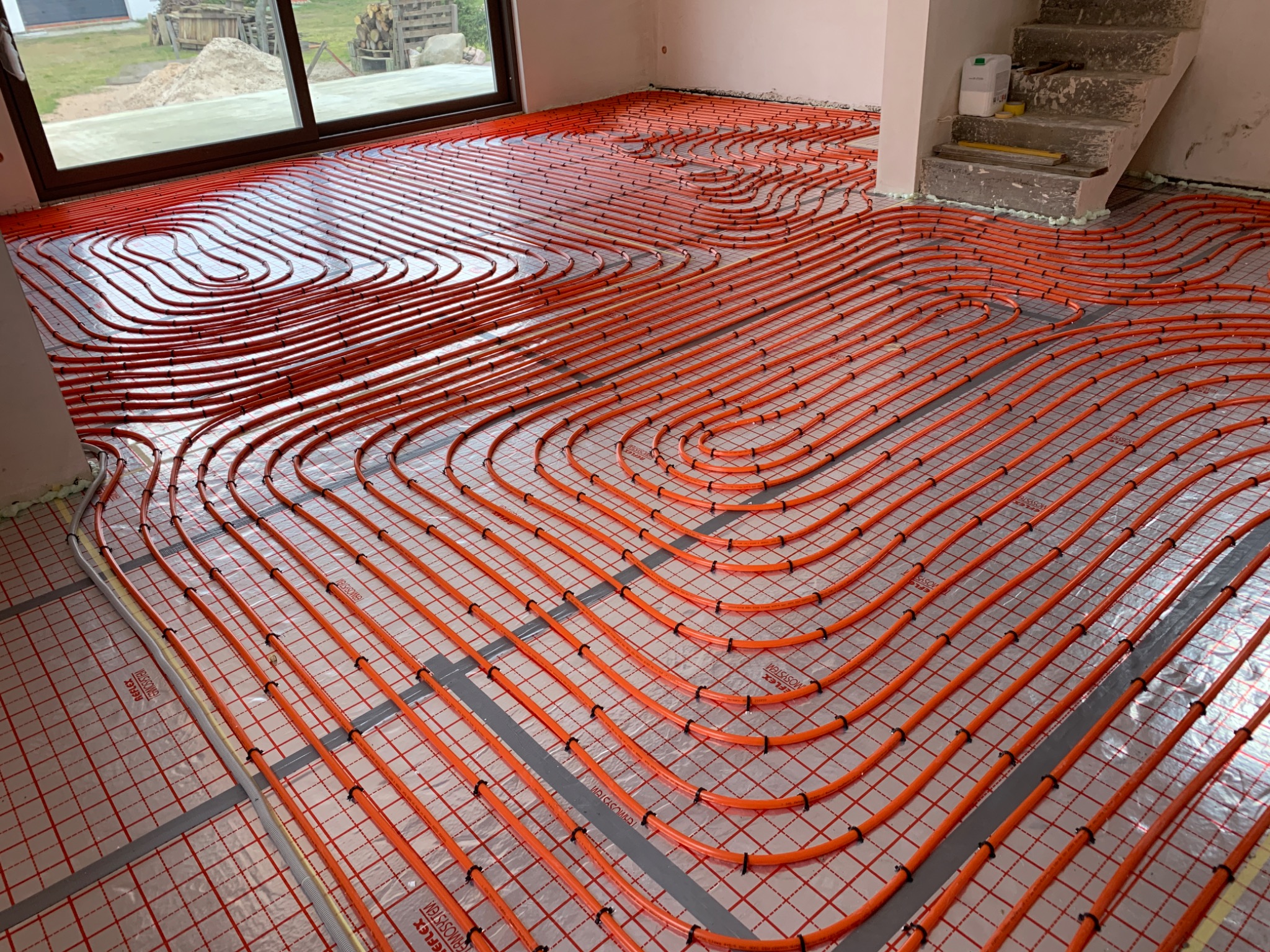 Pomarańczowe rury ogrzewania podłogowego ułożone na srebrnej folii refleksyjnej z czarnymi mocowaniami, widoczne fragmenty ścian i schodów w surowym stanie.