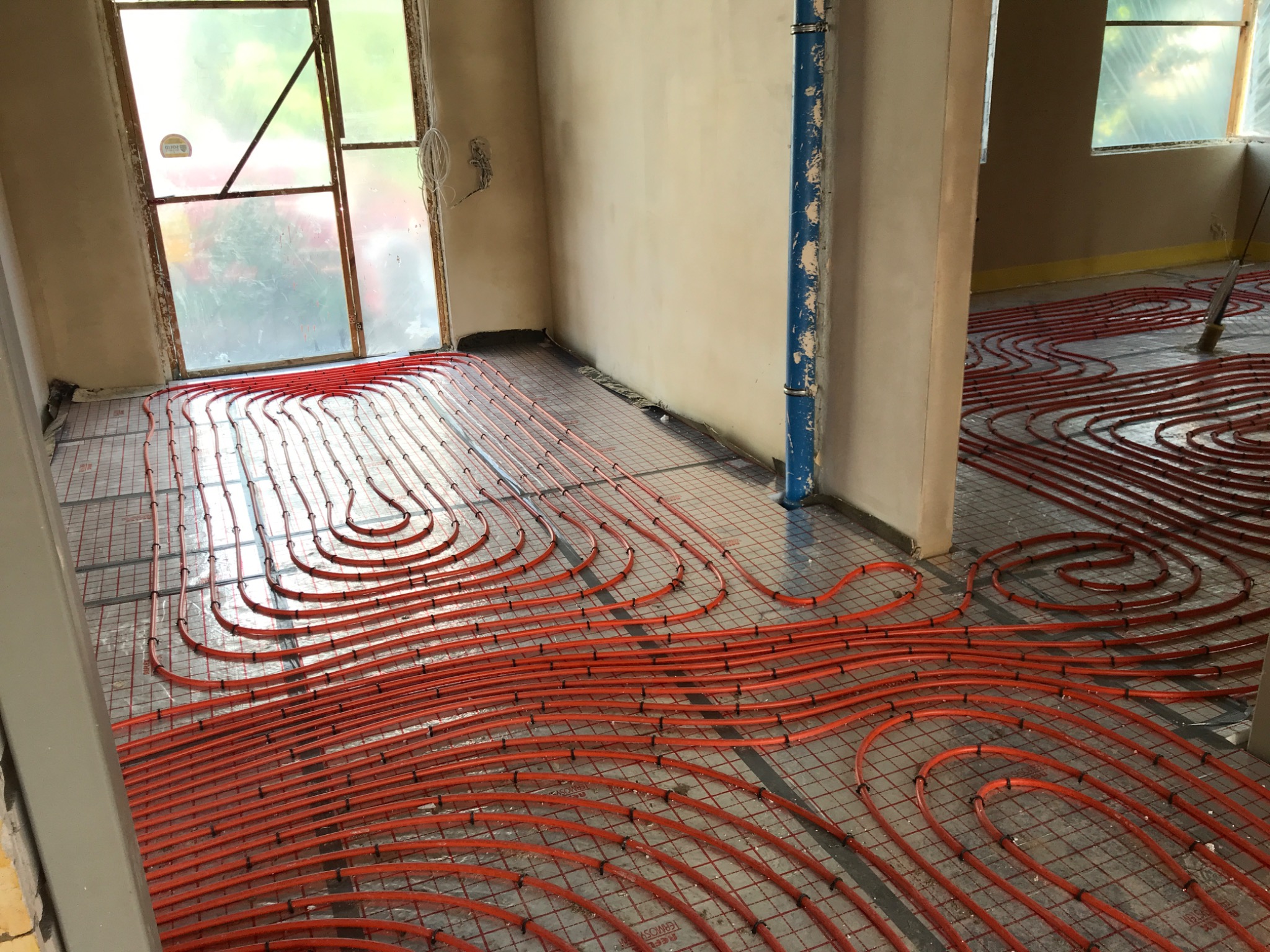 Pomieszczenie w trakcie instalacji ogrzewania podłogowego: czerwone rurki ułożone na siatce, widoczne okna i surowe ściany.