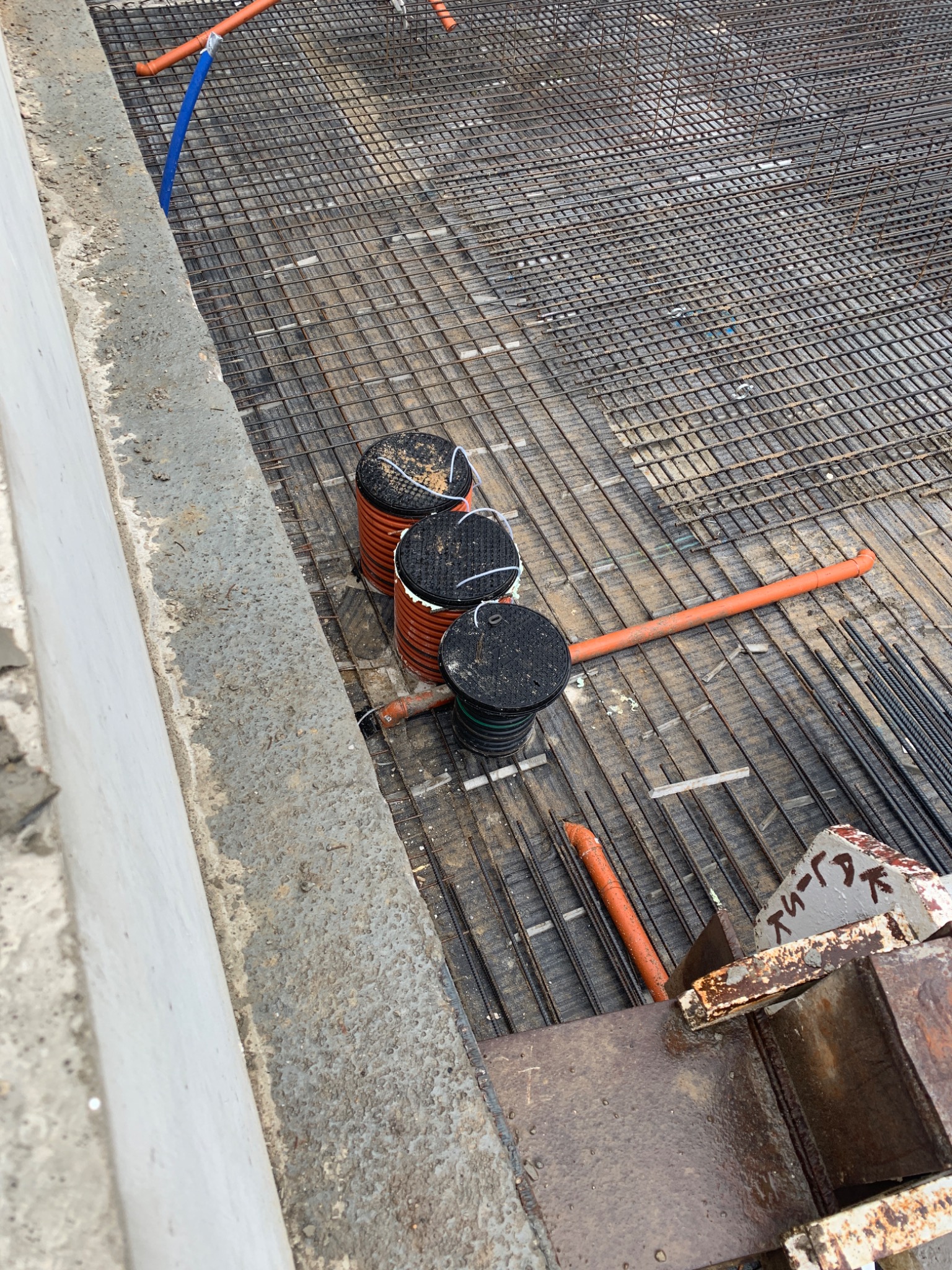 Widok z góry na plac budowy z siatką zbrojeniową, pomarańczowymi rurami i trzema czarnymi studzienkami kanalizacyjnymi z pomarańczowymi pierścieniami, znajdującymi się w pobliżu betonowej ściany.
