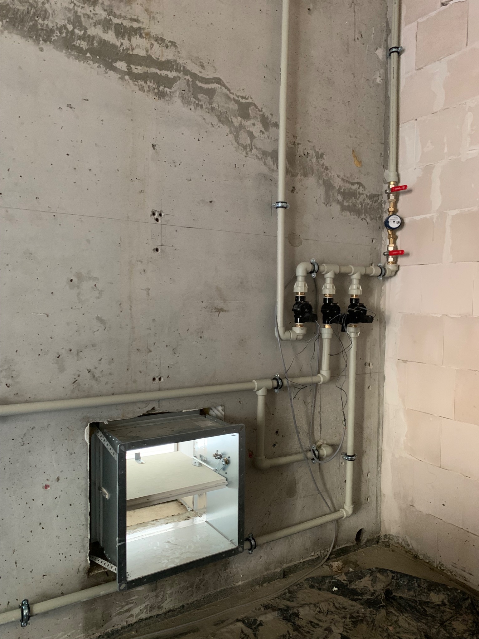 Szare rury instalacyjne z zaworami i licznikiem, zamontowane na surowej ścianie z betonu komórkowego i betonu, z widoczną otwartą szafką rewizyjną z półkami, w trakcie prac instalacyjnych.