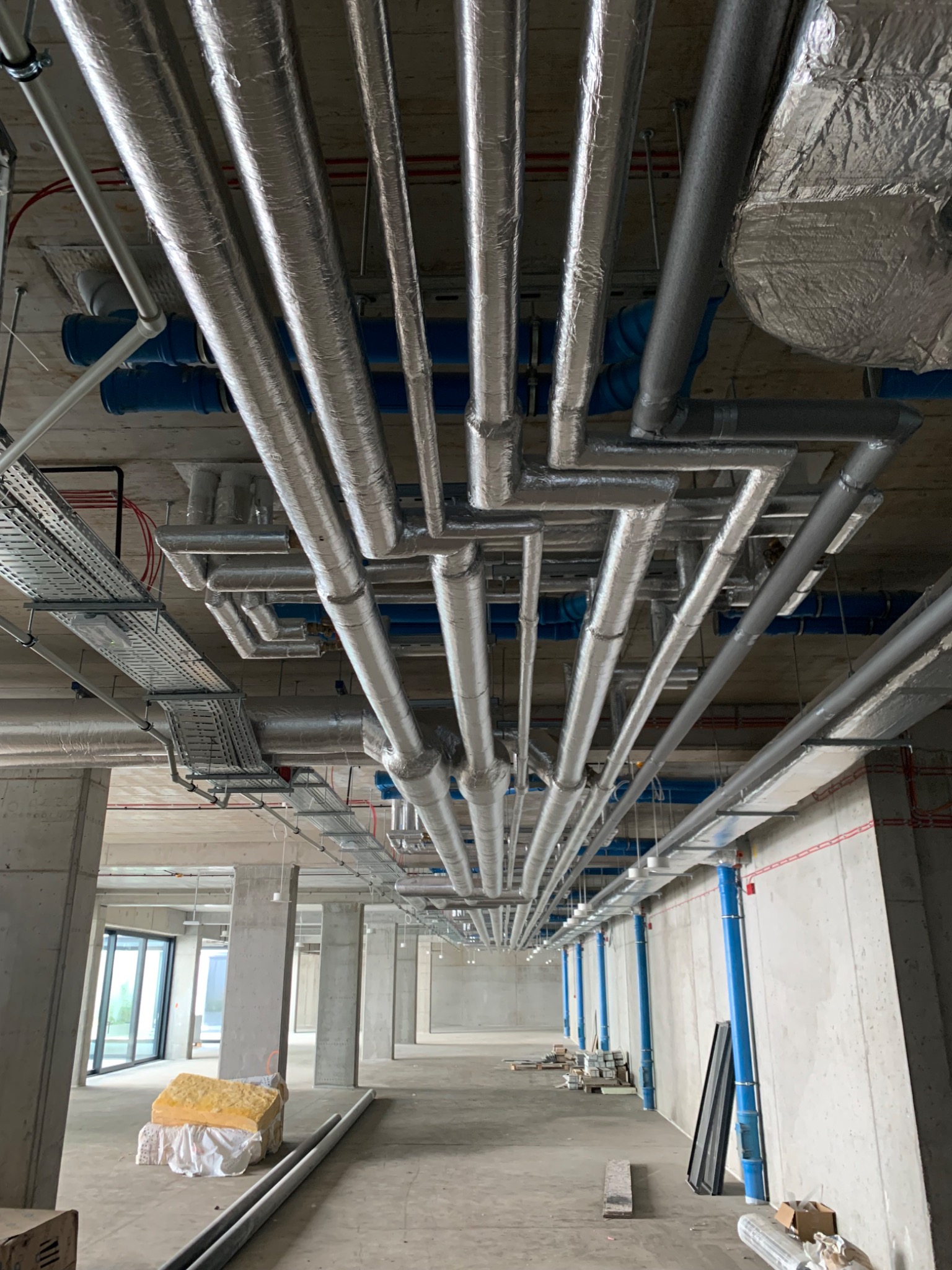 Widok z dołu na system rur wentylacyjnych i instalacyjnych pod sufitem w surowym stanie, z widoczną izolacją i elementami mocującymi w nowo budowanym obiekcie.