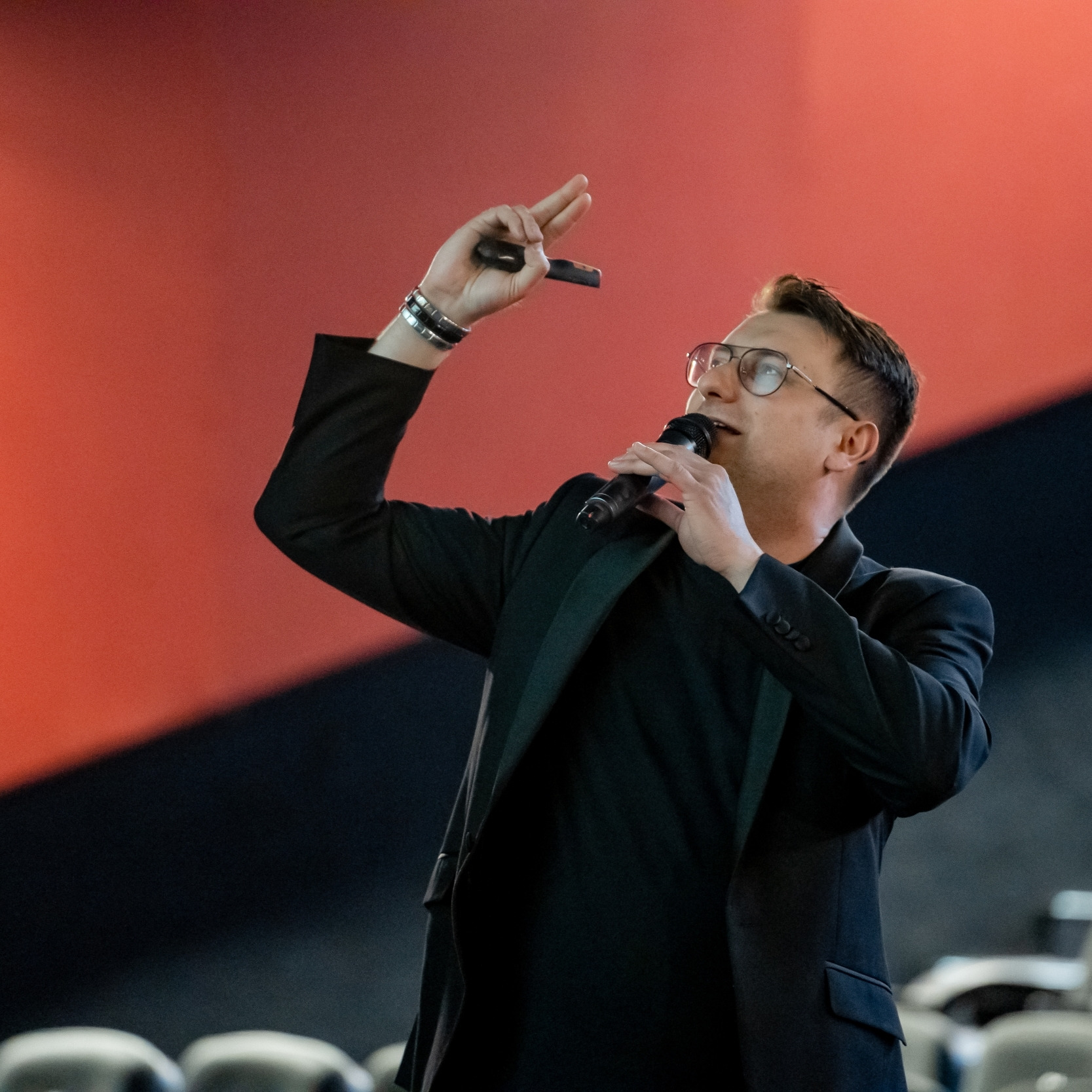 Mężczyzna w okularach i czarnym garniturze, trzymający mikrofon i gestykulujący ręką z bransoletkami na tle czerwono-czarnej ściany.