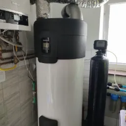 Biała pompa ciepła Buderus z czarnym panelem i srebrną rurą wentylacyjną, obok czarny filtr wody z wyświetlaczem, na tle beżowych płytek i okna.