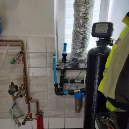 Zestaw instalacji wodnej z czarnym filtrem kolumnowym, rurami miedzianymi i plastikowymi, widocznymi narzędziami i fragmentem okna w pomieszczeniu z kafelkowymi ścianami.