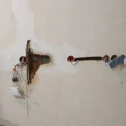 Ściana w trakcie prac elektrycznych: wycięte otwory pod puszki, wystające przewody, zamontowany przełącznik światła, widoczne ślady po gipsowaniu.