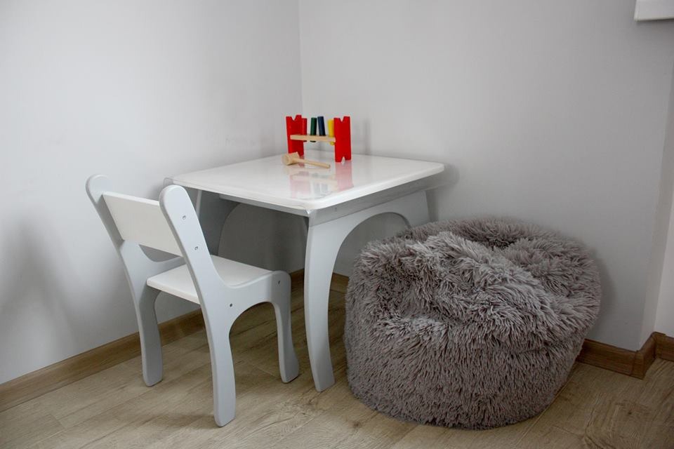 Jasnoszary stolik i krzesełko dla dziecka z drewnianym cymbałkiem i młoteczkiem na blacie, obok puszysty szary puf w pokoju.