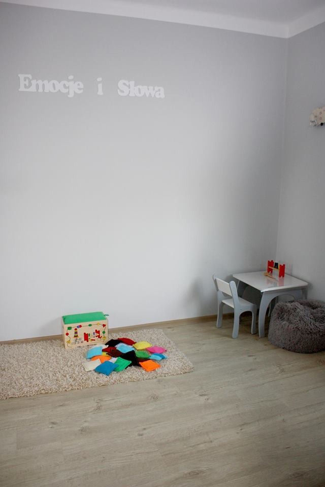 Jasne wnętrze gabinetu logopedycznego w Ciechanowie z napisem 'Emocje i Słowa' na ścianie, stolikiem i krzesłem dla dzieci oraz pudełkiem z kolorowymi woreczkami na dywanie.