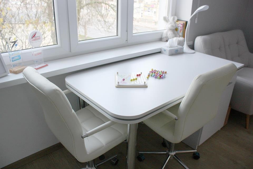 Jasny gabinet z białym biurkiem, krzesłami na kółkach i zabawkami edukacyjnymi na blacie, widok zza krzesła.