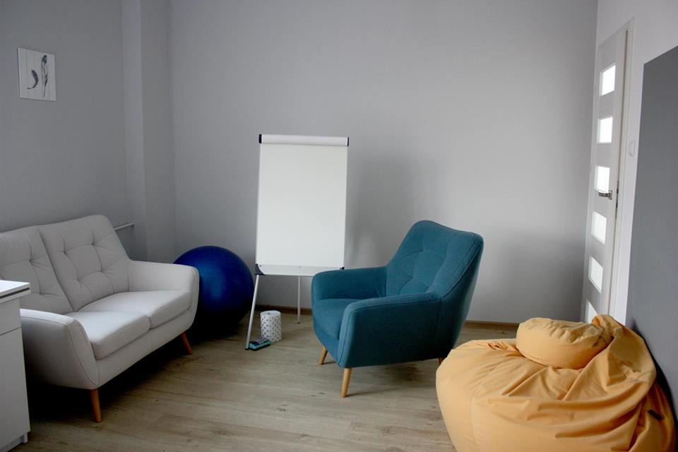 Jasne wnętrze gabinetu z białym biurkiem, kanapą, niebieskim fotelem, workiem sako, piłką do ćwiczeń i tablicą flipchart