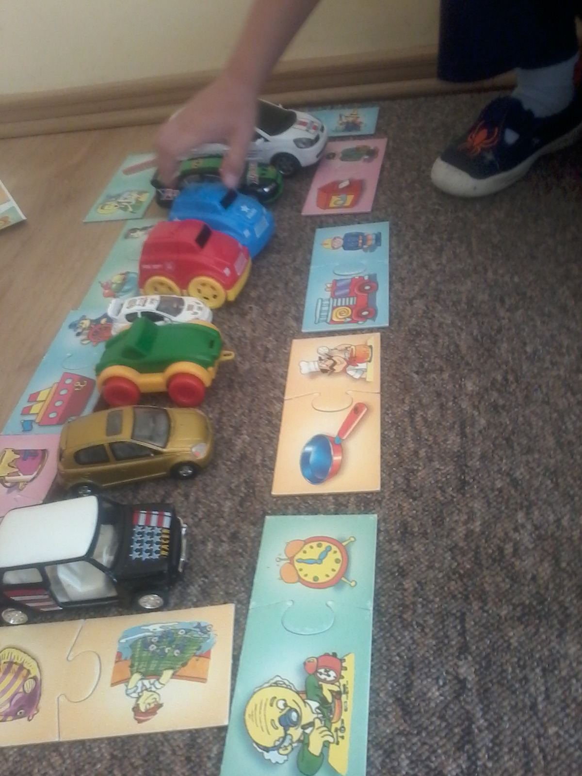 Dziecko układa kolorowe, plastikowe autka na kartach puzzli z obrazkami, na brązowym dywanie.