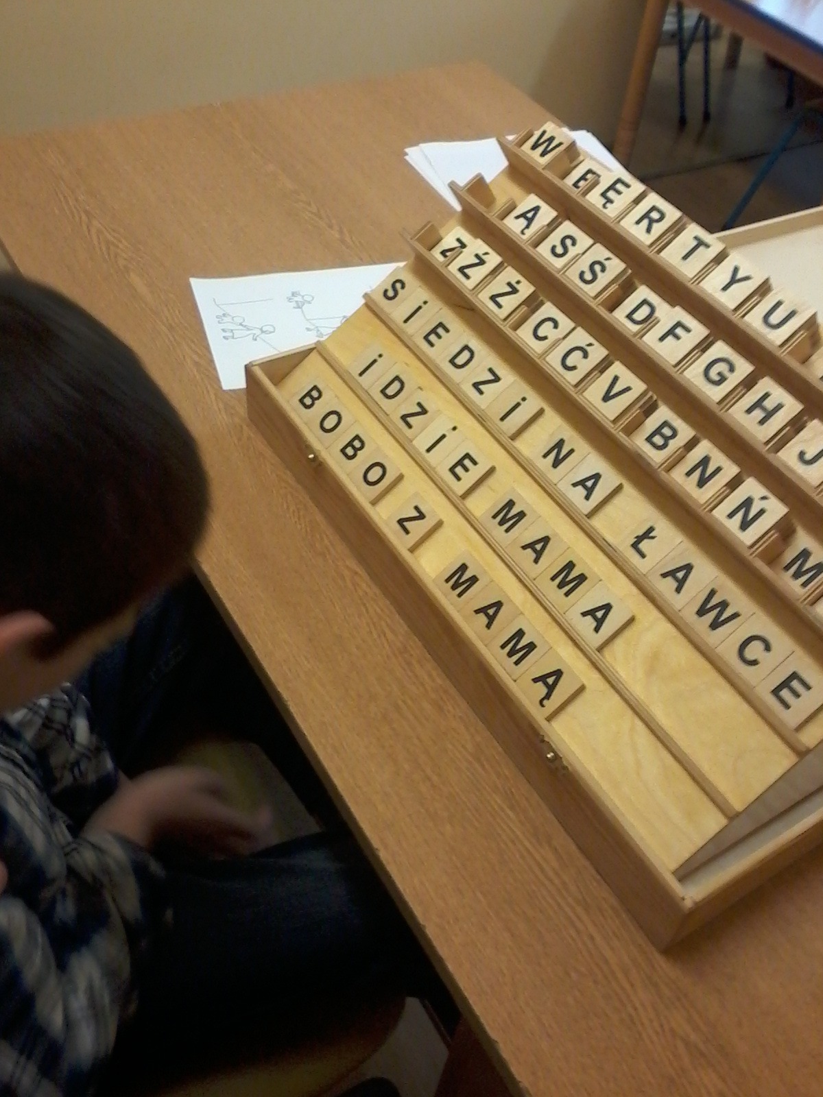 Chłopiec siedzi przy stole z drewnianymi klockami z literami, układając słowa 'BOBO Z MAMA', 'IDZIE NA ŁAWCE', 'SIEDZI' oraz alfabet w drewnianej kasecie.