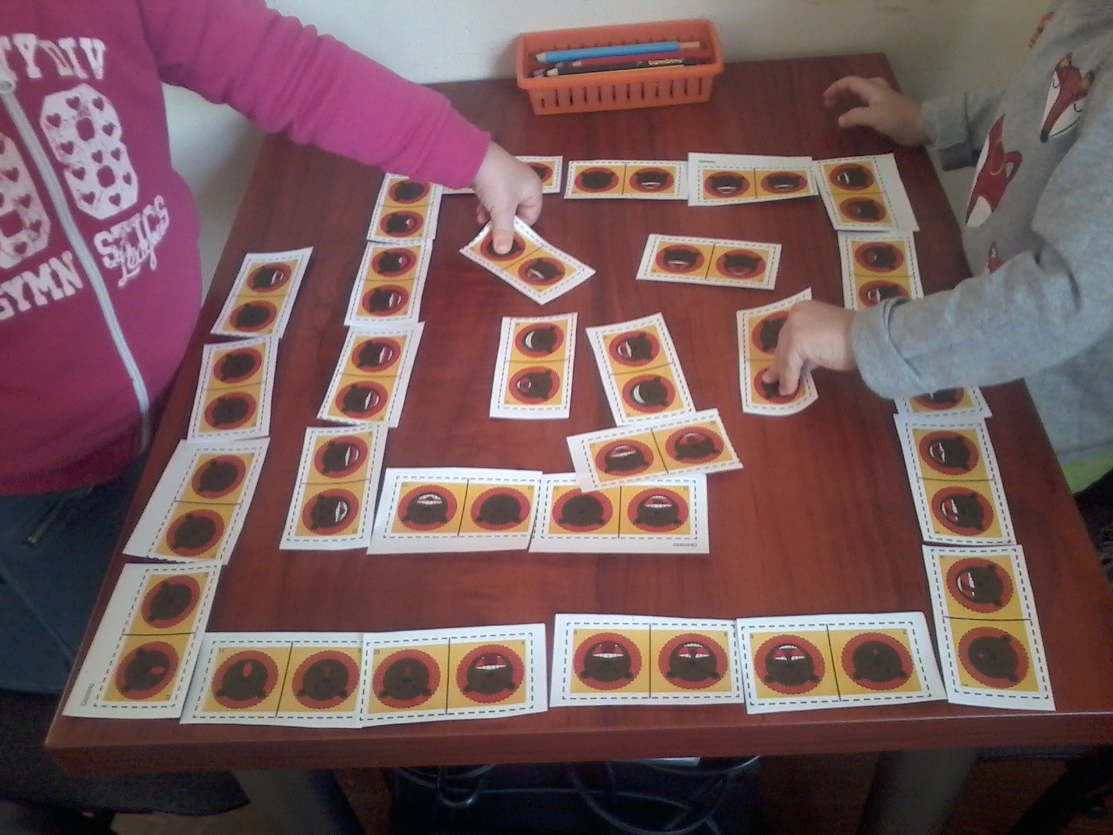 Dzieci grają w grę edukacyjną z obrazkami ust i języka na drewnianym stole, pomoce logopedyczne w pomarańczowym pojemniku w tle.