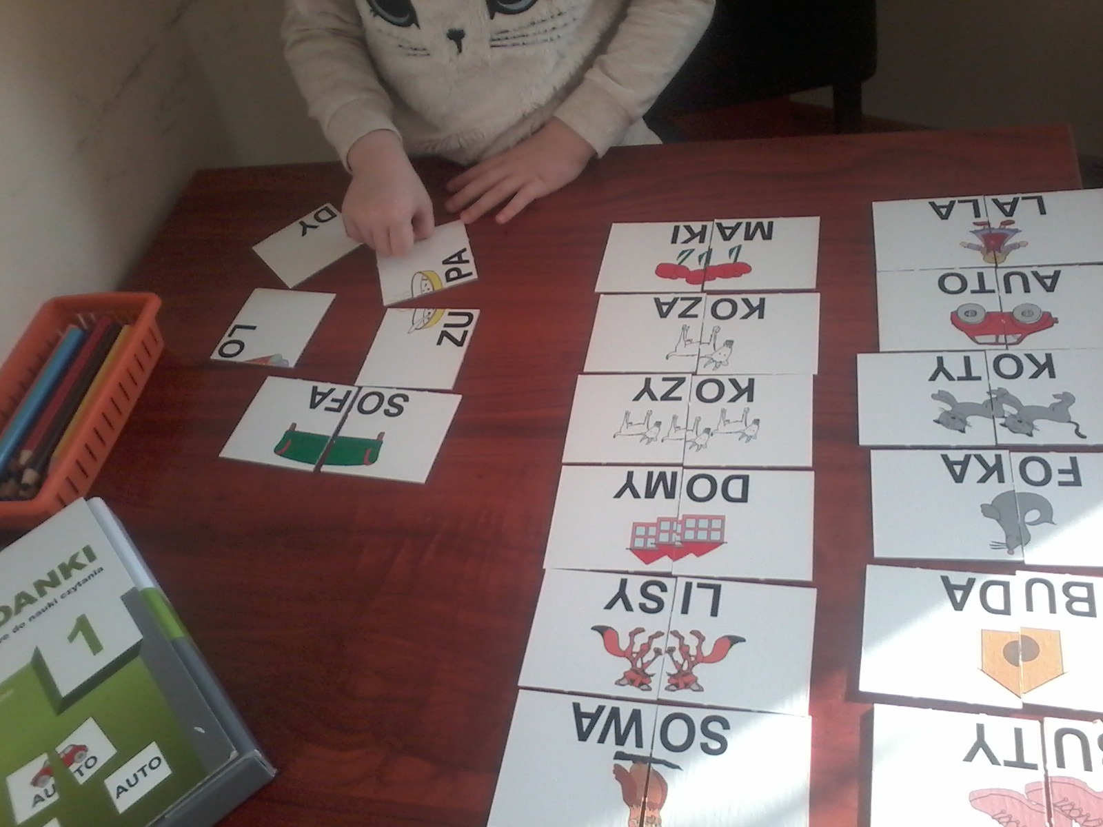 Dziecko w swetrze z motywem kota układa puzzle edukacyjne z obrazkami i słowami na drewnianym stole, obok pudełko z kredkami i książka do nauki czytania.