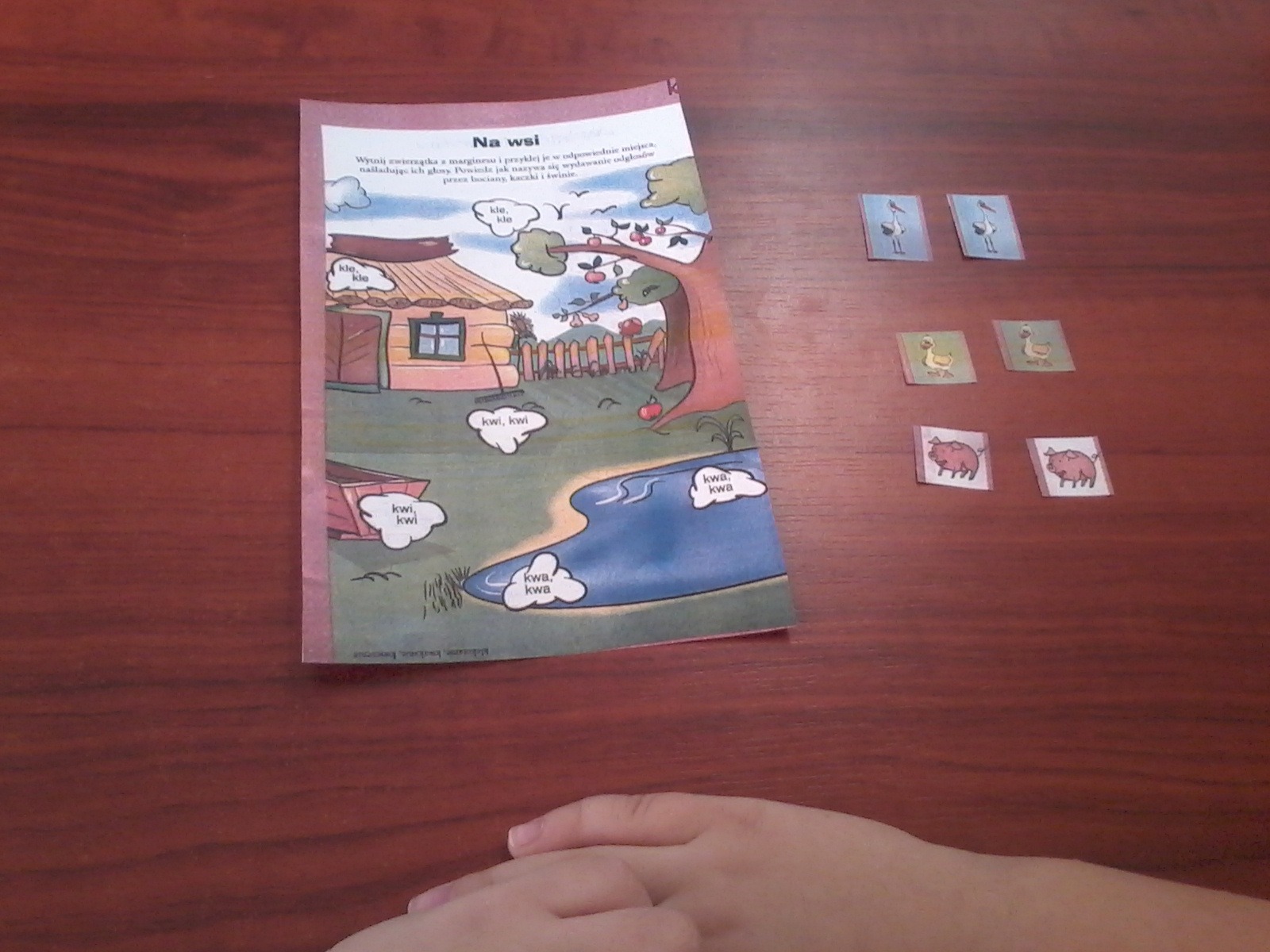 Książka z obrazkami wiejskiego krajobrazu i naklejkami zwierząt na drewnianym stole, ilustracja do nauki odgłosów zwierząt.