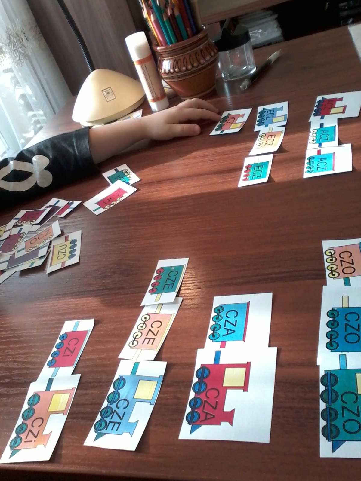 Dziecko układa na drewnianym stole karty z sylabami i obrazkami lokomotyw, obok klej w sztyfcie i pojemnik z kredkami.